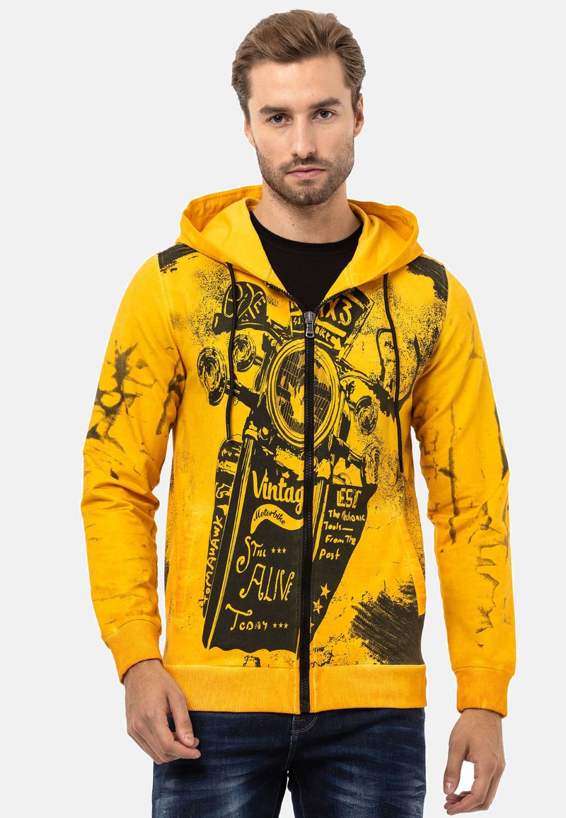 Haut à capuche zippé jaune avec un graphique de moto vintage noir, capuche à cordon et poches avant. Le tissu a une texture lisse et légèrement usée.