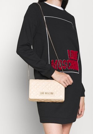 Prošívaná béžová kabelka s řetízkovým popruhem, zdobená zlatou logovou plaketu a měkkou koženou texturou, nošená k černé mikině.