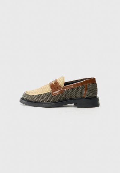 Textured green and tan slip-on shoes with a woven upper, brown leather strap, and smooth black sole. Accented with a patterned detail on the strap.