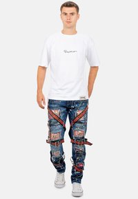 T-shirt blanc avec écriture noire, associé à un jean bleu déchiré ornés de taches de peinture multicolores et d'accents rouges avec des patchs.