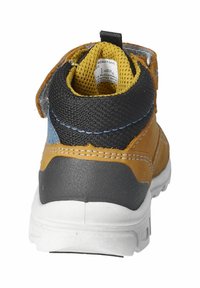 Zapato de cuero marrón con acentos de textil negro, forro de malla amarilla y suela de goma blanca; cuenta con una correa de Velcro y un detalle de costura azul.