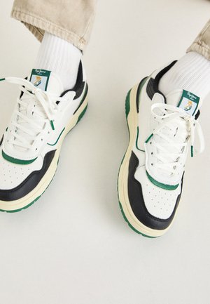 Baskets blanches et noires avec des accents verts, bout rond, tige en cuir perforé, design à lacets et semelle en caoutchouc présentées avec des chaussettes blanches.