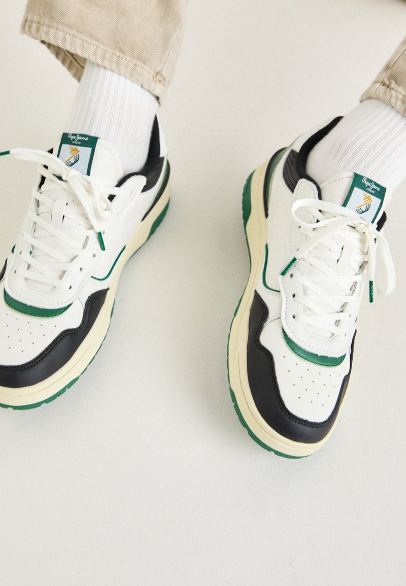 White and black sneakers with green accents, round toe, perforated leather upper, lace-up design, and rubber sole displayed with white socks.