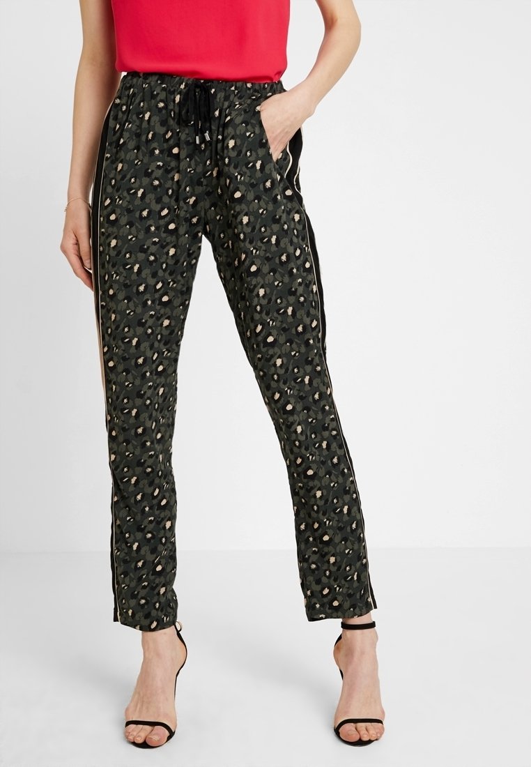 Femme portant un pantalon noir à imprimé léopard avec des bandes latérales, associé à un haut rouge et des talons ouverts noirs, debout avec une main dans la poche.