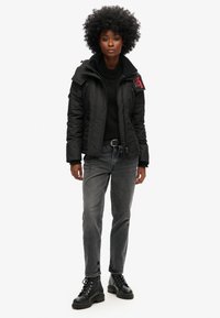 Sort polstret jakke med høj krave, lynlåsåbning og rød logo detalje. Båret sammen med en sort sweater, grå jeans og sorte snørebåndsstøvler.