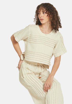 Ženska z kodrastimi lasmi nosi kremno teksturirano kratko majico z rokavi in ustrezne hlače z vrvico, sedi in pozira ob enostavnem ozadju.