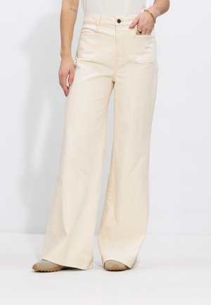 Persoon draagt een crème-kleurige broek met hoge taille en brede pijpen, voorzien van opgestikte voorzakken, staat met één hand in de zak en zichtbare beige schoenen.