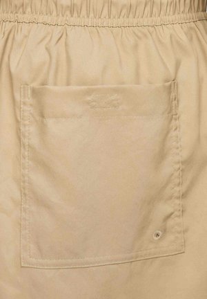 Beige stof med en glat tekstur, der har en elastisk talje og en stor klaplomme på bagsiden med synlige syninger.