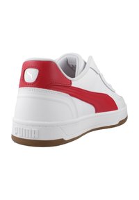 Zapatilla blanca con una franja de cuero rojo y acento en el talón. Suela de goma plana con fondo texturizado marrón. Presenta un logo de Puma en el talón.