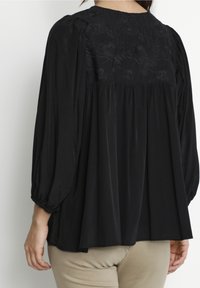 Svart blus med en texturerad blommig broderad rygg, lös passform och ballongärmar. Mjuk tyg faller lätt över kroppen.