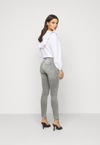 Blusa branca com detalhes em babados, combinada com jeans skinny cinzentos. Os jeans apresentam uma bainha desfiada e uma silhueta justa, complementados por mules de salto pretos.