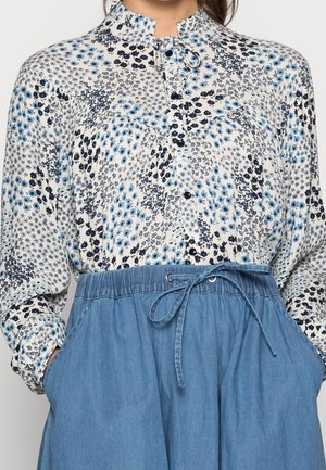 Bluse mit floralem Muster in Blau und Marineblau auf hellem Hintergrund, mit Kragen, Knöpfen und einem lockeren Design, kombiniert mit einem blauen Kordelzugrock.