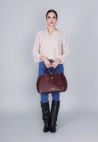 Ruskea teksturoitu käsilaukku kultaisella soljella, yhdistettynä beigeen puseroon, sinisiin farkkuihin ja mustiin polvimittaisiin saappaisiin. Yksinkertainen ja tyylikäs muotoilu.