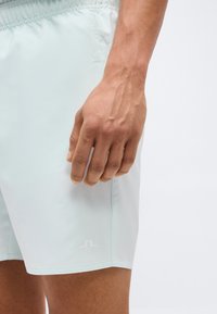 Δεξί χέρι ακουμπισμένο σε ανοιχτόχρωμο σορτς με ελαστική μέση και μικρό λογότυπο κοντά στο τελείωμα.