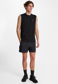 En ung man står klädd i en ärmlös svart topp, svarta shorts, svarta strumpor i ankellängd och svarta sneakers mot en vit bakgrund.