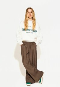 Bijela dukserica s zelenim natpisom, smeđe široke hlače i zelene tenisice. Outfit se odlikuje ležernim krojem i teksturiranim materijalom.