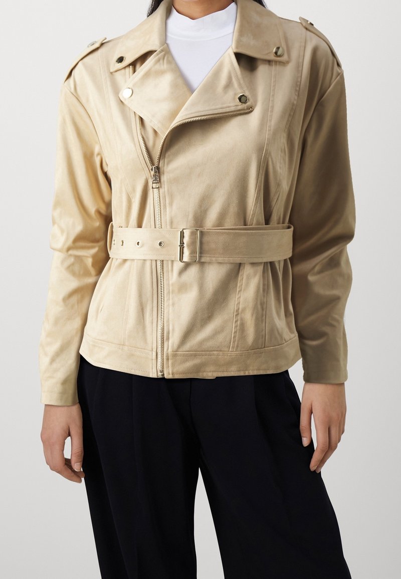 Veste beige ceinturée avec fermeture éclair et boutons-pression portée sur un haut blanc, associée à un pantalon noir ample sur une personne debout.