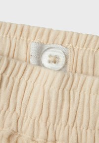 Beige ribbad tyg med en texturerad yta, med en klar knapp och en vit knappöglan upptill. Mjukt, stretchigt material.