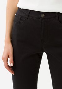 Nahaufnahme einer Hand, die schwarze Hose mit einem Knopf und Taschen hält, während ein weißes Oberteil getragen wird, vor einem neutralen Hintergrund.