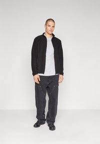 Svart fleecejakke med høy krage, grå t-skjorte under, svarte baggy-jeans og svart fottøy. Enkel design og nøytrale farger.