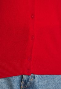 Rote Strickjacke mit geripptem Saum und sechs passenden Knöpfen entlang der Vorderseite. Kombiniert mit leicht blauer Jeansstoff am unteren Ende.