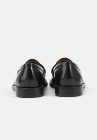Zwarte leren schoenen met een gepolijste afwerking, voorzien van een ronde neus, middenhoge hak en dubbele gesps. Het geheel heeft een gladde textuur.