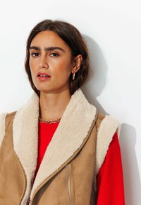 Gilet en daim synthétique beige avec un col en fausse fourrure crème, doté d'une fermeture éclair et d'une texture douce, porté par-dessus un haut à manches longues rouge.