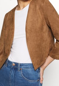 Veste courte en daim marron avec manches trois-quarts, associée à un haut blanc côtelé et un jean en denim bleu. Texture lisse et coupe ajustée.