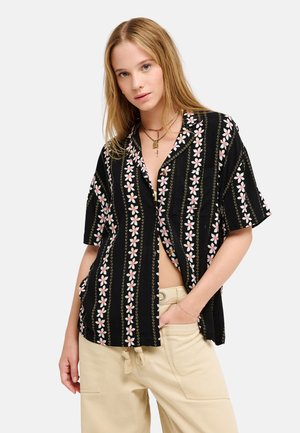 Jeune femme aux longs cheveux blonds portant un t-shirt noir à manches courtes avec des rayures florales roses et un pantalon beige, posant avec une main dans la poche.