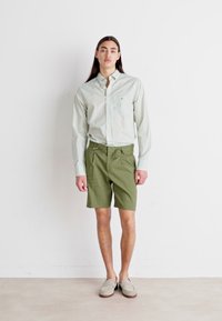 Persoană tânără cu păr lung și închis la culoare, îmbrăcată într-o cămașă button-up cu carouri verzi deschise, pantaloni scurți verzi măslinii și încălțăminte slip-on bej, stând în fața unui perete alb.