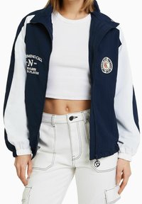 Veste zippée bleu marine et blanche avec un col montant, des poignets élastiques et un patch logo à l'avant. Portée sur un crop top blanc et un pantalon clair.