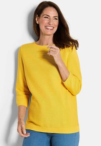Gelbes Sweatshirt mit Dreiviertelärmeln, Rundhalsausschnitt, gerippter Textur und einfarbig, kombiniert mit blauen Jeans.