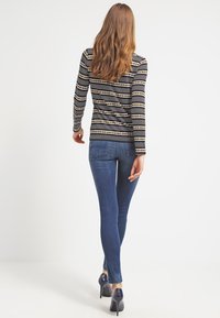 Gestreiftes langärmeliges Oberteil in Schwarz und Beige, kombiniert mit blauen engen Jeans und High Heels, von hinten betrachtet.