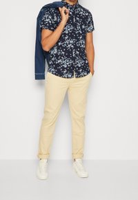 Camicetta a maniche corte con stampa floreale blu navy, pantaloni beige e sneakers bianchi; giacca di jeans appoggiata sulla spalla; vestibilità casual e tessuto leggero.