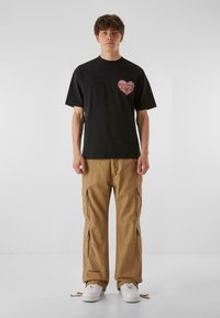 Svart bomullströja med hjärt-grafik som läser "Heartbreak Hotel", som matchas med khaki cargobyxor med sidofickor och vita sneakers.