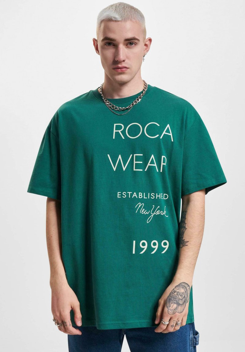 Grøn oversized t-shirt med hvid tekst, der står "ROCA WEAR ESTABLISHED New York 1999." Bomuldsstof med korte ærmer.