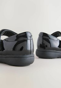 Chaussures en cuir verni noir avec une finition lisse, bout rond, bande Velcro et semelle en caoutchouc noir texturé. La vue arrière montre des détails de couture.