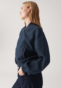 Giacca bomber navy con colletto alto, zip frontale e tasche laterali. Presenta maniche a palloncino e un tessuto liscio e testurizzato.