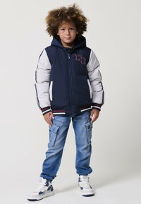 Dieťa s kučeravými vlasmi oblečené v námorníckej a bielej hoodie, modrých cargo džínsach a bielych teniskách stojí čelom.