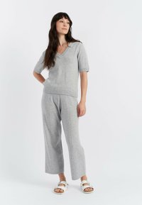 Maglione grigio a maglia con maniche corte e scollo a V, abbinato a pantaloni grigi a gamba larga, caratterizzati da dettagli delle cuciture. Il modello indossa sandali bianchi.