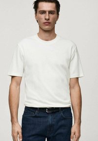 Tricou alb din bumbac, cu guler rotund și mâneci scurte, purtat împreună cu blugi din denim albastru închis și o curea neagră. Textură fină, design simplu.