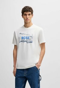Hvid kortærmet t-shirt med et blåt "BOSS" logo og et blåt abstrakt kortdesign på forsiden, parret med blå denimbukser.