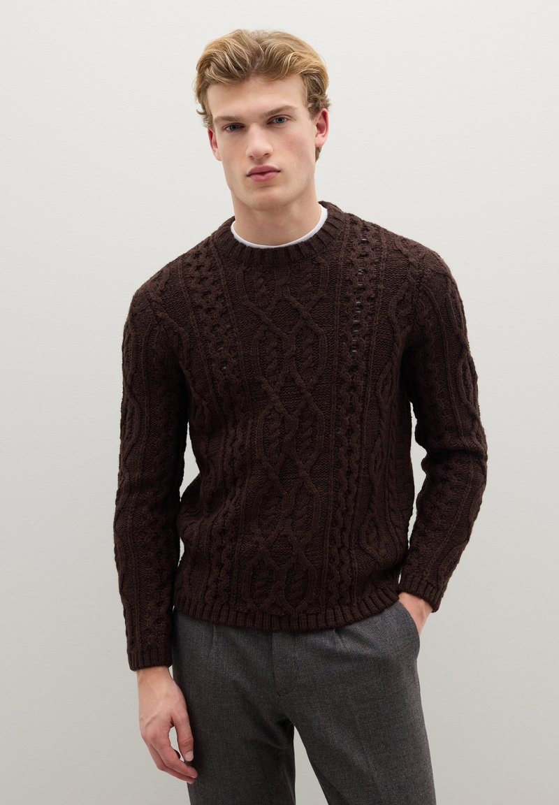 Pull marron en tricot avec un motif torsadé, col rond, poignets et ourlet côtelés. Porté avec un pantalon gris sur mesure.