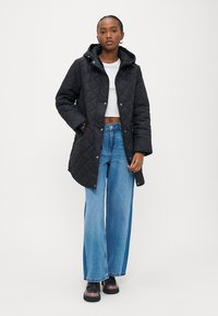 Ženska, oblečena v črn prešit plašč s kapuco, beli kratki zgornji del, široke modre kavbojke in črne platformne čevlje, stoji pred belim ozadjem.