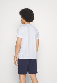 Camiseta atlética blanca de manga corta con cuello redondo, textura suave y pantalones cortos oscuros en contraste, vista desde atrás.