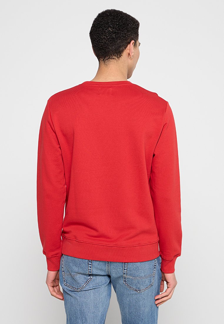 Mann in einem schlichten roten Langarmpullover und blauen Jeans, der mit dem Rücken zu einer einfarbigen hellen Hintergrund steht.