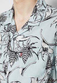 Camicia azzurro chiaro a maniche corte con stampa di ananas nero e motivi floreali, con taschino frontale e bottoni bianchi, realizzata in tessuto liscio.