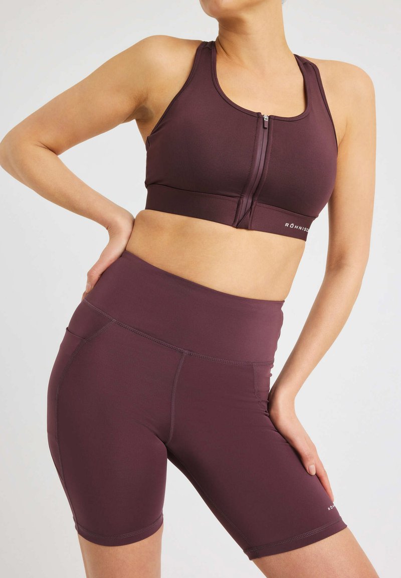 Burgundy urheilBra, jossa on vetoketju edessä ja leveät hihnat, yhdistettynä yhteensopiviin korkeavyötäröisiin shortseihin. Sileä kangas ja minimalistiset muotoiluyksityiskohdat.