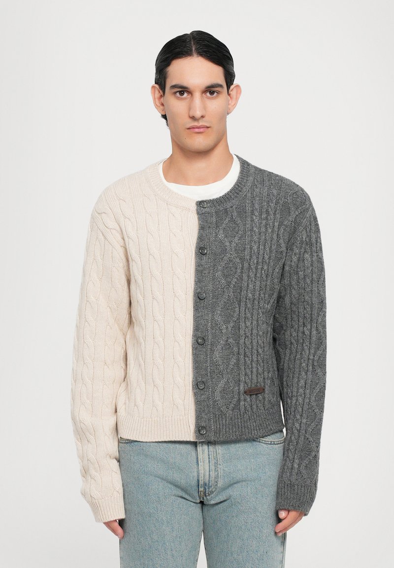 Stickad kofta med en delad design, en sida i kräm och en mörkgrå sida. Texturerade flätmönster och knäppning framtill.