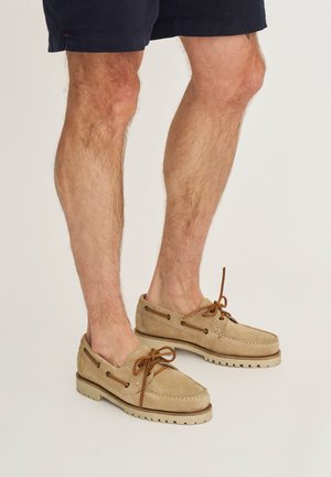 Beige suède herensloffen met bruine leren veters, gedragen bij marineblauwe shorts, vanaf halverwege de kuit tot de voeten afgebeeld tegen een effen achtergrond.